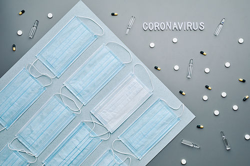 Top down composition of medical masks with coronavirus word and medication laid out in pattern over grey background, copy space