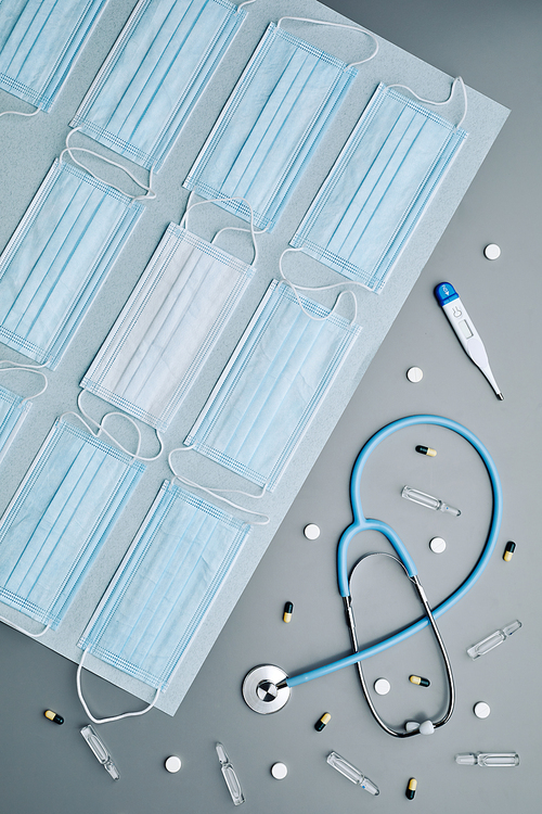 Top down graphic composition of medical masks with stethoscope and thermometer laid out over grey background, copy space