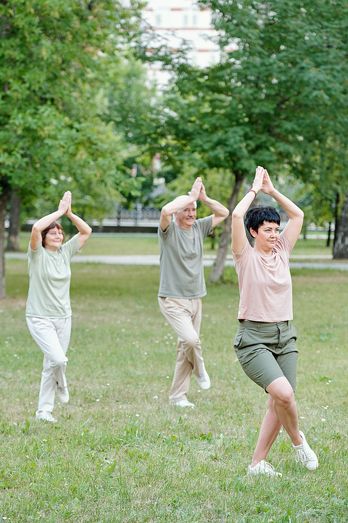 Qigong coach standing with crossed legs and teaching seniors to do balance exercise in park