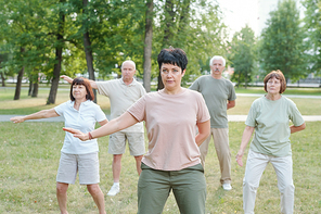 Mature brunette instructor moving arm aside and practicing qigong with senior students in park