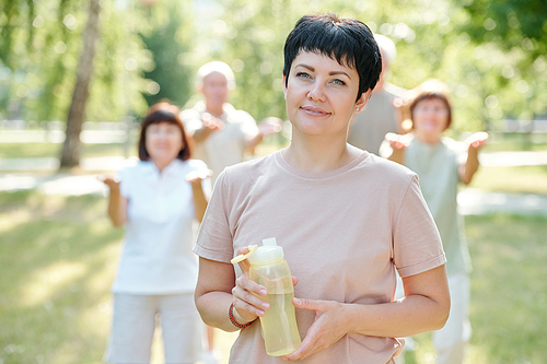 Portrait of smiling brunette fitness coach with short hair holding water bottle against senior people doing exercise