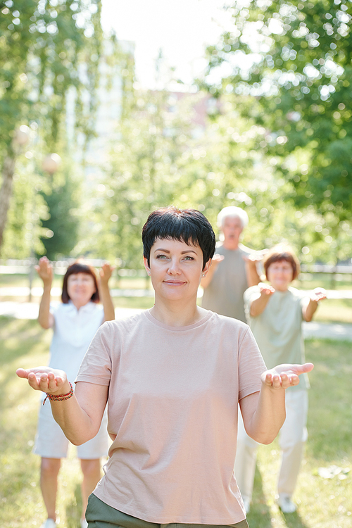 Portrait of smiling qigong teacher showing palms against her students in summer park