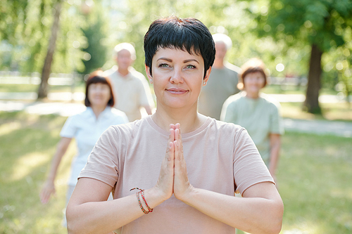 Portrait of content serene mature yoga instructor making Namaste gesture while standing in front of her students in park