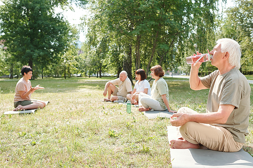 Senior man with mustache sitting with crossed legs on mat in park and drinking water after yoga practice