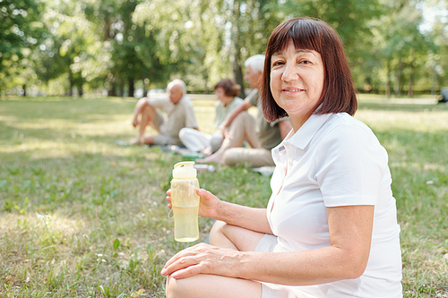 Portrait of content mature lady in white tshirt sitting on grass at outdoor training and drinking water