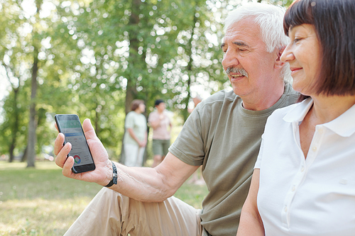 Modern senior man with mustache analyzing day activity with wife using mobile app in park