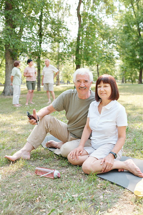 Portrait of positive active senior couple sitting with smartphone on yoga mat in park