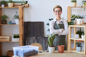 Waist up portrait of smiling young woman wearing apron standing by wooden table with potted plants and soil ready for potting, home gardening concept, copy space
