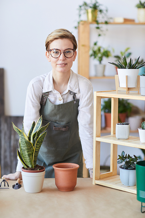 Vertical portrait of modern young woman wearing apron standing by wooden table with potted plants and soil ready for potting, home gardening concept