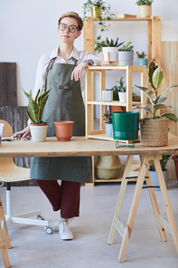 Vertical full length portrait of modern young woman wearing apron standing by wooden table with potted plants and soil ready for potting, home gardening concept