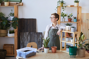 Waist up portrait of modern young woman wearing apron standing by wooden table with potted plants and soil ready for potting, home gardening concept, copy space