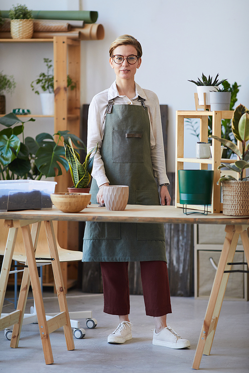 Vertical full length portrait of modern young woman smiling at camera while standing by wooden table with plants and soil ready for potting, home gardening concept