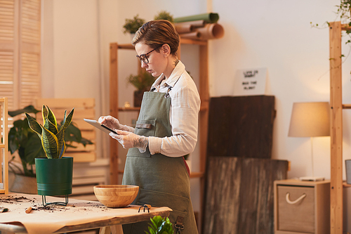 Side view portrait of modern young woman using digital tablet while potting houseplants indoors, copy space