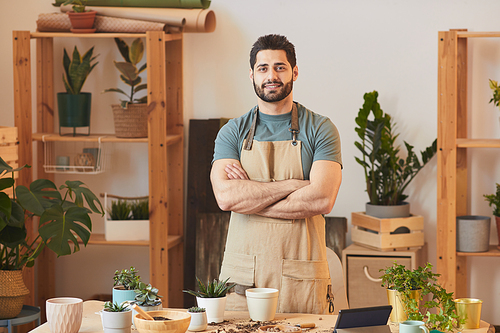 Waist up portrait of handsome bearded man wearing apron and  while standing by table with houseplants and gardening tools, copy space