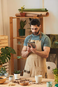 Vertical portrait of handsome bearded man wearing apron and using digital tablet while standing by table with houseplants and gardening tools