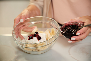 Hands of young woman holding small bowl with blackcurrant jam and a larger one with ingredients of homemade icecream over table