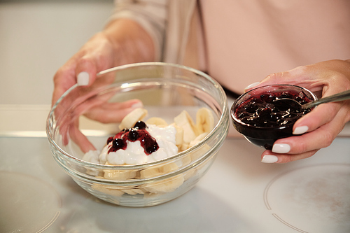 Hands of young woman holding small bowl with blackcurrant jam and a larger one with ingredients of homemade icecream over table