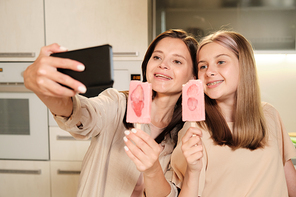 Two young cheerful females with long hair making selfie in the kitchen while eating homemade eskimo icecream with strawberry slices