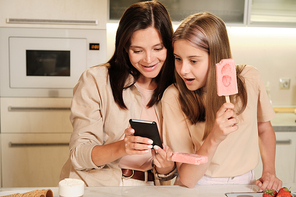 Young mother with smartphone showing her surprised teenage daughter curious online video while both having homemade icecream