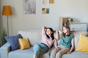 Young mother and daughter relaxing on couch in living-room against wall with pantings, lamp, green domestic plant and some furniture