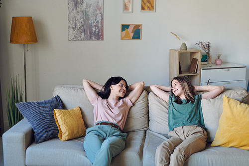 cheerful and relaxed mother and daughter in casualwear looking at each other while sitting on soft couch and chatting in home