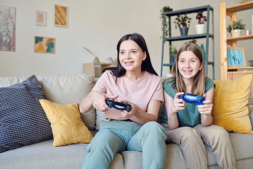 Young smiling mother and cute teenage girl with joysticks pressing buttons during video game while sitting on couch against shelves by wall