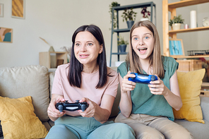 Excited blond teenage girl with joystick sitting on couch next to happy mother while both playing video game in front of tv set at home