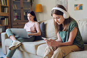 Serious teenager girl choosing song via smartphone app and listening to music in headphones while her mother working with laptop