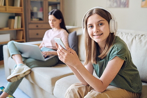 Portrait of positive modern teenager in wireless headphones using smartphone while her busy mother working on laptop