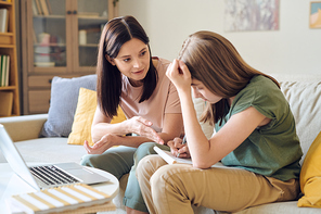 Young confident brunette woman in casualwear helping her teenage daughter with homework while both sitting in front of laptop