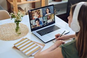Over shoulder view of student girl in headphones sitting at table and participating in online conference in student group