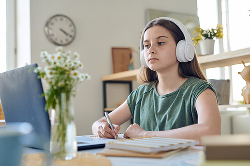 Diligent serious teenage schoolgirl in headphones listening to her teacher explanation while sitting in front of laptop during remote lesson