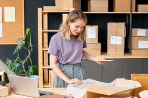 Pretty young blond woman with mobile gadget standing by table and looking at folded winterwear in open box before packing the order
