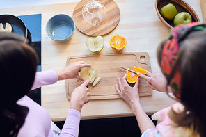 Above view of unrecognizable women cutting apple and orange on wooden board while making fruit salad in kitchen
