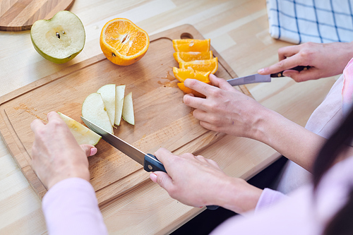 Hands of two young contemporary females with knives slicing fresh oranges and apples on wooden board while preparing breakfast