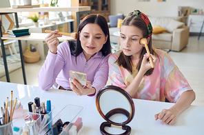Young brunette woman showing her cute teenage daughter how to apply powder or blush on face while both looking in mirrors at home