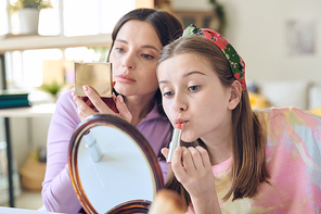 Cute teenage girl with long blond hair applying pink lipstick and looking in mirror on background of her mother with open compact powder