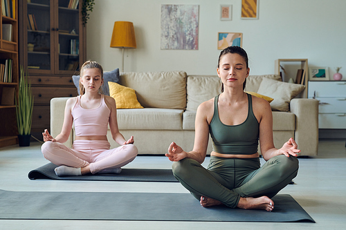 Relaxed brunette female and her teenage daughter in tracksuits sitting on mats in pose of lotus while taking yoga course for beginners at home