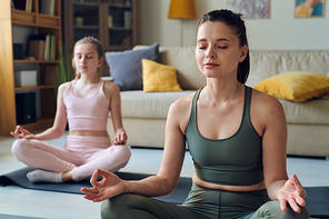 Calm middle-aged mother meditating with closed eyes together with daughter at home