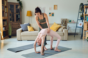 young active woman in grey sportswear helping her teenage daughter in pink tracksuit during backbend exercise on mat in home