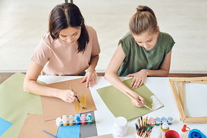 Young woman and her teenage daughter with paintbrushes painting with watercolors or gouache on paper while sitting by desk at home
