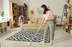 Young contemporary woman in casualwear washing floor of living-room while standing by carpet on background of her daughter cleaning sofa