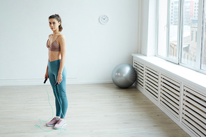 Minimal full length portrait of fit young woman holding skipping rope and  during home workout in white room, copy space