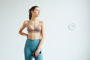 Minimal waist up portrait of fit young woman holding skipping rope and looking away while enjoying fitness workout standing against white wall with analog clock, copy space