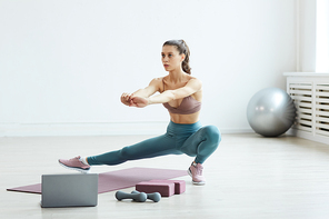 Minimal full length portrait of fit young woman stretching legs while enjoying sports workout at home in white room, copy space