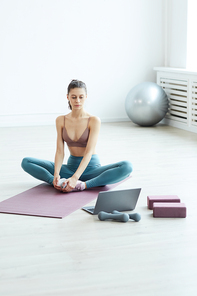 Minimal full length portrait of fit young woman warming up and stretching while watching workout video via laptop at home, copy space