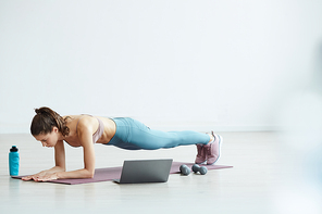 Minimal full length portrait of fit young woman doing plank exercise while watching workout video via laptop at home, copy space