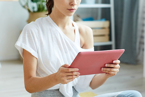 Cropped portrait of contemporary young woman holding digital tablet while sitting on yoga mat at home, copy space