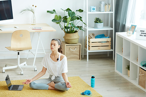 Full length portrait of contemporary young woman meditating at home while sitting in lotus position during yoga workout, copy space