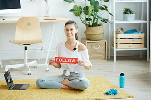 Full length portrait of smiling young woman holding FOLLOW sign while recording workout video at home and , copy space
